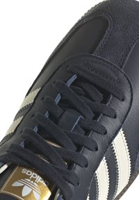 Ingrandimento di una sneaker Adidas blu scuro con lacci neri, strisce bianche sui lati e un'etichetta con logo dorato sulla linguetta.