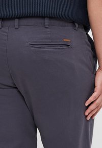 Dark gray trousers made of textured fabric, featuring a back pocket with a horizontal opening and a small leather brand tag.
