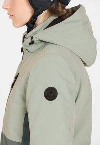 Hellgrüne Skijacke mit Kapuze, strukturiertem Stoff und einem schwarzen runden Logoschild auf dem linken Ärmel. Sichtbare Reißverschlusstasche.
