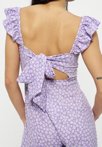 Femme portant une robe sans manches violette à fleurs avec des bretelles volantées et un grand nœud attaché dans le bas du dos.