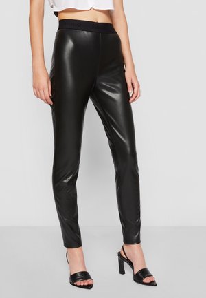 Femme portant des leggings en simili cuir noir avec une ceinture élastique et des sandales noires à talons hauts, debout devant un fond uni.