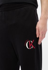 Μαύρες φόρμες από μαλακό ύφασμα, με ελαστική μέση και λογότυπο Calvin Klein σε κόκκινο και λευκό χρώμα στο αριστερό μηρό.