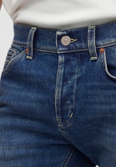 Gros plan sur la partie médiane portant un jean denim bleu avec bouton et passants de ceinture, associé à un haut blanc rentré.
