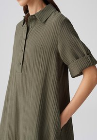 Vestito camicia verde oliva realizzato in un tessuto texturizzato a coste. Presenta un colletto, un'apertura con bottoni, maniche arrotolate e tasche laterali.