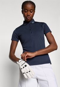 Polo navy com gola, botões e detalhes de logótipo; o modelo usa uma luva de golfe branca com detalhes pretos, combinada com calças de cor clara.