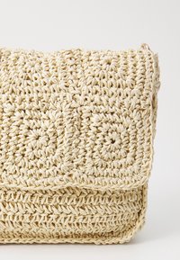 Bolso cruzado de paja tejida beige con cierre de solapa, que presenta un patrón circular de crochet floral y un sutil anillo dorado en el lateral.