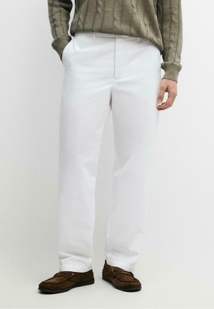 Mann trägt weiße gerade geschnittene Hose, braune Wildleder-Loafer und einen olivgrünen Zopfpullover mit Händen in den Taschen.