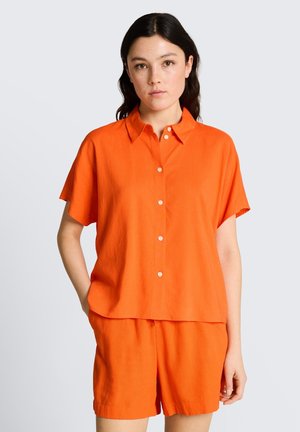 Femme portant une chemise orange à manches courtes boutonnée et un short orange assorti, debout avec une main dans la poche.