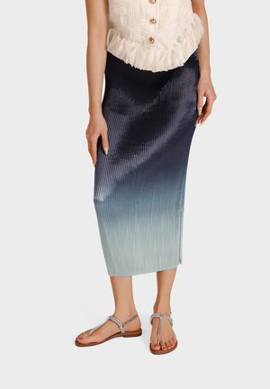 Donna che indossa una gonna midi plissettata ombré dal blu scuro al blu chiaro con sandali piatti argentati con cinturini su sfondo bianco.