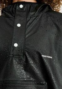 Svart vattentät jacka med hög krage, tryckknappar och synliga vattendroppar. Har en framficka och en logotyp på den nedre sidan.