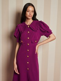 Vestido fúcsia com gola larga e frisada, mangas curtas com volume e detalhes em botões dourados; conta com dois bolsos frontais e uma cintura ajustada.