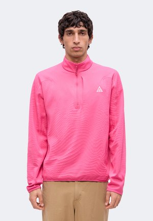Junger Mann mit kurzen lockigen dunklen Haaren, der einen knallpinkfarbenen Viertel-Reißverschluss-Pullover und beige Hosen trägt, steht vor einem schlichten Hintergrund.