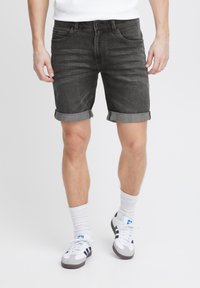 Ungdommelig voksen iført sorte rullede denimshorts, hvide crew sokker og hvide sneakers med sorte striber, står op ad en ensfarvet baggrund.