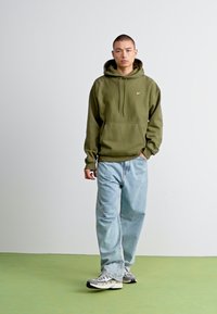 Olivengrøn hættetrøje med snore og en frontlomme, parret med løse lyseblå jeans og grå sneakers. Enkel, afslappet design.