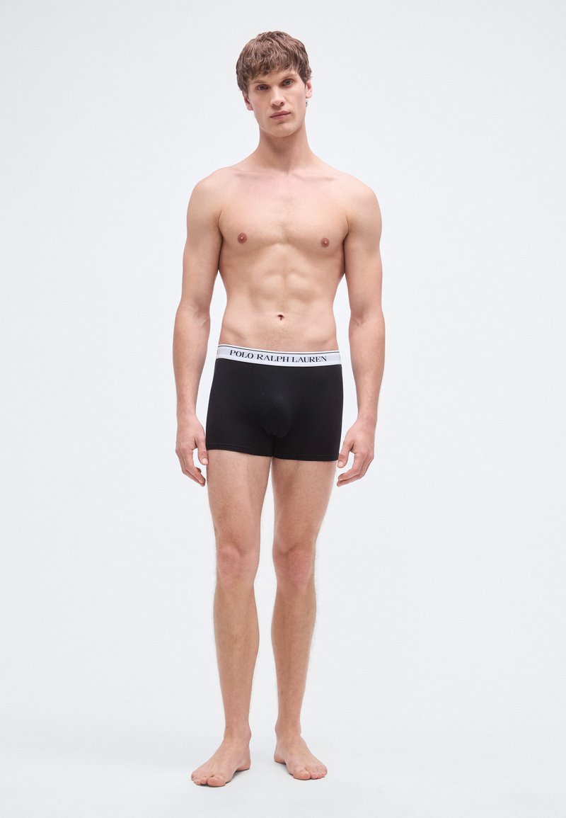 Cuecas boxer de algodão pretas com uma cintura branca que exibe "POLO RALPH LAUREN" em letras pretas. O modelo está descalço e de frente.