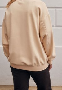 Beige oversized sweatshirt met verlaagde schouders en geribbelde boorden, gecombineerd met zwarte broek. Zachte textuur, minimale naden, casual ontwerp.