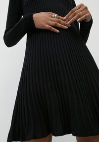 Robe noire côtelée à manches longues, silhouette évasée, tissu texturé et encolure arrondie. Les détails notables incluent une coupe ajustée à la taille.