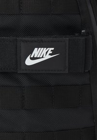 Juodas „Nike“ kuprinės detalės, pasižyminčios tekstūruota medžiaga, dirželiais su tinklu ir stačiakampiu logotipo ženklu su baltu tekstu bei „swoosh“ simboliu.
