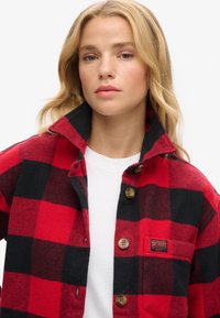 Camicia di flanella a scacchi rossa e nera con colletto, chiusura con bottoni e taschino sul petto con una patch del logo del brand. Texture morbida.