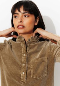 Chemise en velours côtelé marron avec un col à volants, patte de boutonnage à cinq boutons et une petite poche. Présente une texture côtelée verticale et une couleur unie.