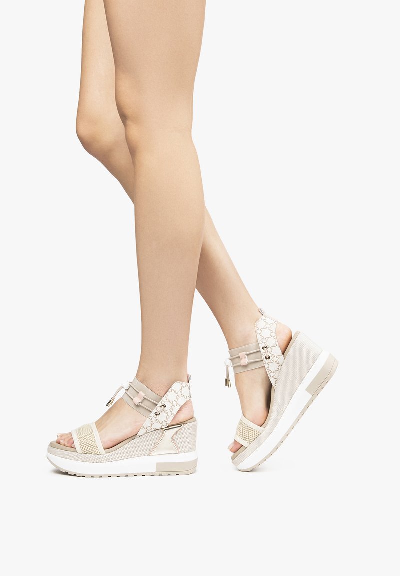 Beige kile-sandaler har en tekstureret mesh-design, kontrasterende stropper, en skrå sål og dekorative hardware-accenter. Ideelle til afslappet brug.