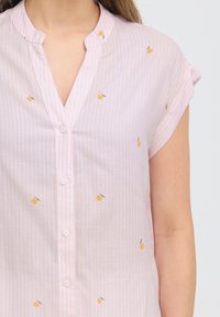 Blouse rose clair à manches courtes avec un petit imprimé de fruits orange, fermeture boutonnée devant et fines rayures verticales.