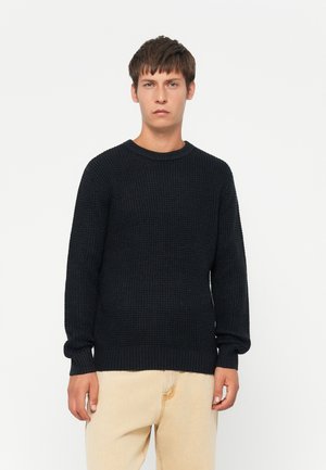 Giovane uomo che indossa un maglione a maglia nera con texture e pantaloni beige chiari, in piedi di fronte a uno sfondo bianco semplice.