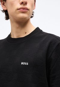 Νεαρός με μαύρο φούτερ με υφή και μικρό λευκό λογότυπο "BOSS" στο στήθος, φωτεινό φόντο.