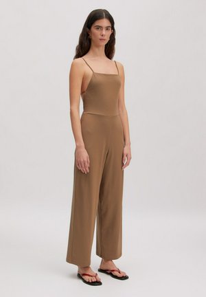 Frau mit langen dunklen Haaren, die einen ärmellosen braunen Overall und schwarze flache Sandalen mit roten Riemen trägt, vor einfarbigem Hintergrund stehend.