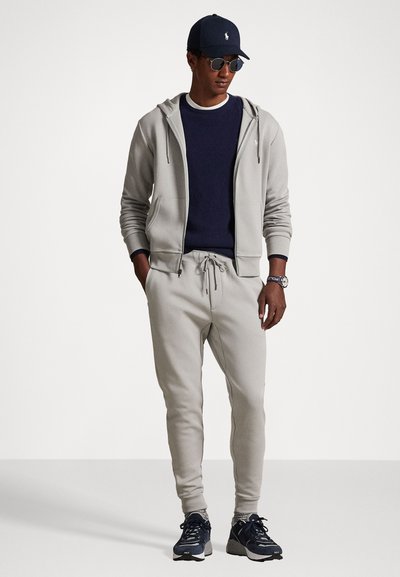 Camisola com fecho éclair cinza claro, sweatshirt azul marinho, calças de fato combinando, ténis pretos e um boné azul marinho com logotipo. Os acessórios incluem óculos de sol e um relógio.