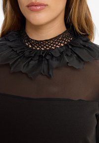 Haut noir avec un col en mesh volanté agrémenté de motifs floraux, associé à un corps entièrement noir. Texture douce avec des panneaux transparents près du décolleté.