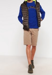 Chaleco acolchado marrón oscuro con capucha sobre una sudadera azul con letras amarillas, combinado con pantalones cortos beige y zapatillas verde oliva.