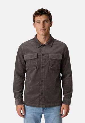 Joven con una camisa de pana gris oscuro con dos bolsillos en el pecho y jeans azul claro, de pie frente a un fondo liso.