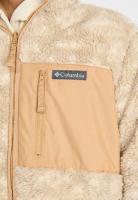 Close-up of beige Columbia fleece jacket with zipper and fabric pocket on chest.
