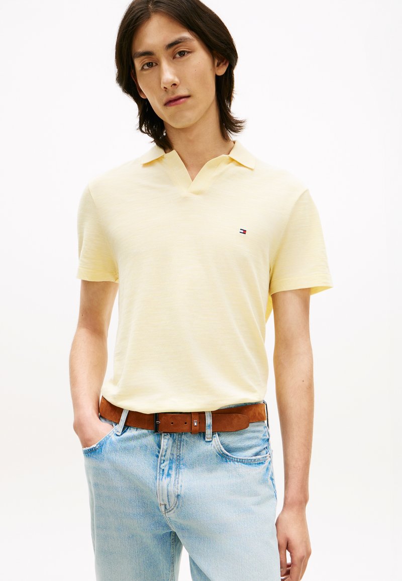 Νεαρός που φοράει απαλό κίτρινο πόλο με κοντά μανίκια και ανοιχτό μπλε τζιν, στέκεται με το ένα χέρι στην τσέπη, μπροστά σε λευκό φόντο.