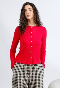 Γυναίκα με μακριά μπούκλια, που φοράει κόκκινο μακρυμάνικο τοπ με κουμπιά και ασπρόμαυρο καρό παντελόνι, στέκεται με τα χέρια στις τσέπες.