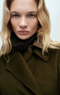 Olive zelený vlnený kabát s veľkým golierom, vrstvený nad tmavým šálom. Modelka má vlnité blond vlasy a neutrálny make-up, ktorý vyjadruje prirodzený výraz.
