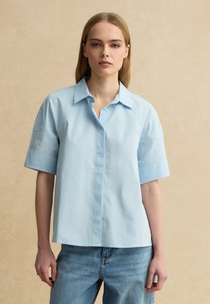 Jonge vrouw met recht blond haar draagt een los lichtblauw shirt met korte mouwen en een blauwe spijkerbroek, staande tegen een beige achtergrond.