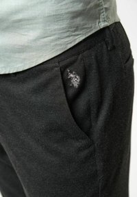 Schwarze Hose aus strukturiertem Stoff mit einem kleinen gestickten Logo in der Nähe der Tasche. Ein hellgrünes Hemd ist teilweise sichtbar.