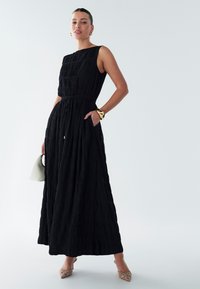 Schwarzes ärmelloses Kleid mit strukturierter Stoffqualität und betonter Taille, ausgestattet mit zwei Seitentaschen. Kombiniert mit einer kleinen weißen Handtasche und verzierten Absätzen.