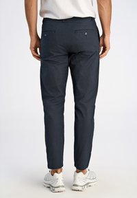 Dunkelblaue Hose mit maßgeschneiderter Passform, ausgestattet mit zwei Gesäßtaschen mit Knopfverschluss und einem glatten, leichten Stoff. Kombiniert mit weißen Sportschuhen.