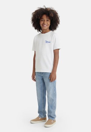 Barn med krøllet hår iført en hvid t-shirt med St. Tropez-logo, lyseblå jeans og beige slip-on sko, smilende og stående.