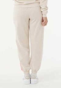 Beige sweatpants met een tapered fit en elastische boorden; voorzien van een contrasterende rode zijstreep; zachte stoftextuur. Draagt met witte sneakers.