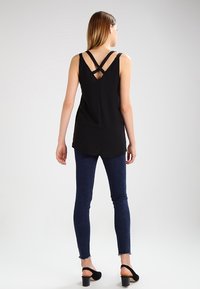 Top negro sin mangas con tirantes en la espalda con cruz, con dobladillo curvado, combinado con jeans skinny de mezclilla oscura y sandalias de tacón negras.