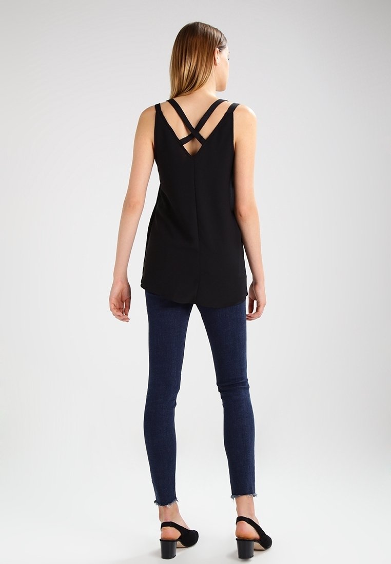 Top negro sin mangas con tirantes en la espalda con cruz, con dobladillo curvado, combinado con jeans skinny de mezclilla oscura y sandalias de tacón negras.