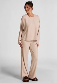 Loszittende beige loungewear set bestaande uit een top met lange mouwen en wijde broekspijpen. Zachte stoftextuur, gecombineerd met bruine, pluizige pantoffels.