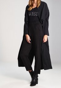 Abrigo largo negro sobre una camiseta negra con texto de lentejuelas, combinado con pantalones sueltos negros y botas de tobillo negras. Tejido suave, ajuste relajado.
