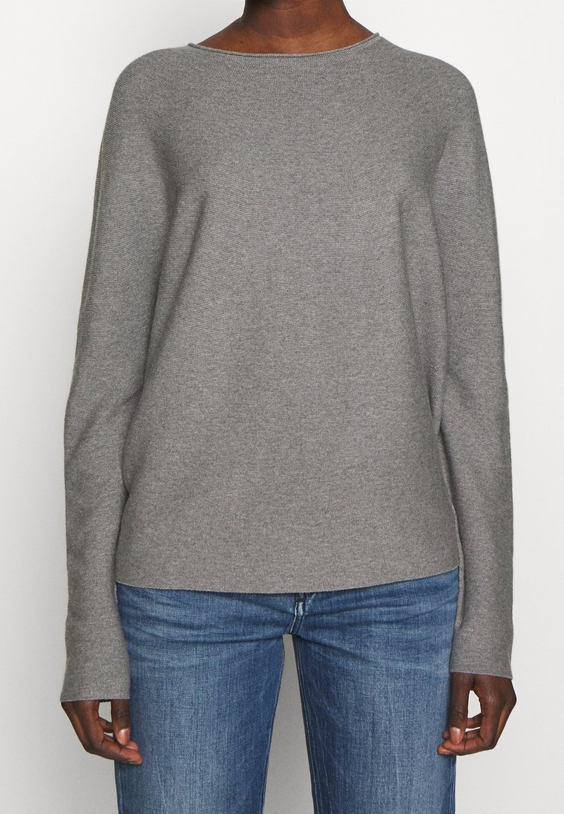 Personne portant un pull en tricot léger gris clair à manches longues et un jean en denim bleu, se tenant devant un fond uni.