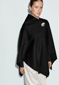 Poncho in lana nera con una texture liscia, caratterizzato da un grande fermaglio argentato e progettato con una silhouette drappeggiata.