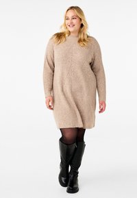 Vestido de punto beige con textura acanalada y patrón geométrico. Mangas largas, corte suelto, combinado con botas negras hasta la rodilla.